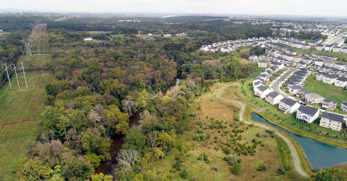 An aerial view of a transmission line cutting through forest on left side of the photo. A large block of forest on either side of goose creek running through the middle of the photo. A suburban housing development can be seen on the right side of the image.