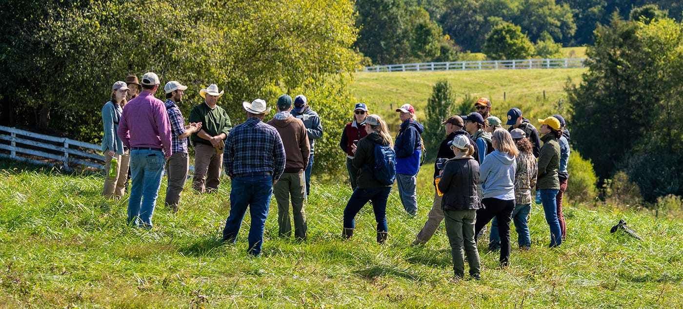 a group of about 20 stands in a farm field listening to a presenter