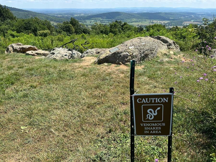 grassy overlook looking out over landscape of open space and farmland with a brown metal sign in the foreground that says "caution venomous snakes in area"