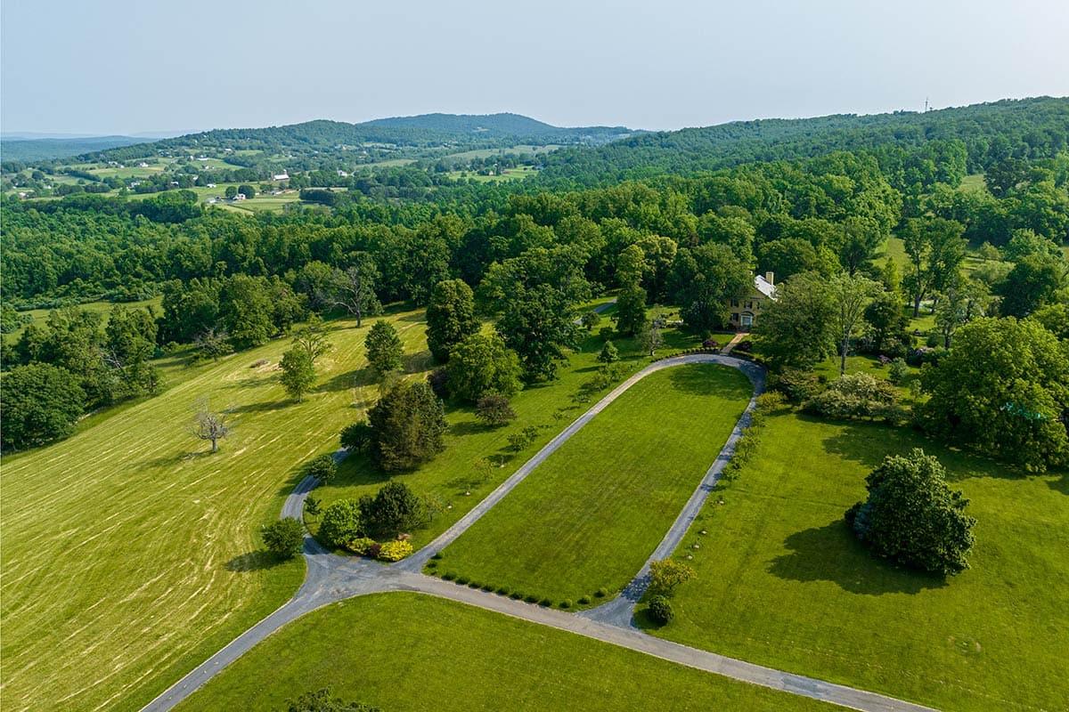 aerial image of big historic house with grassy fields in the foreground and forest in the background with mountains in the distance